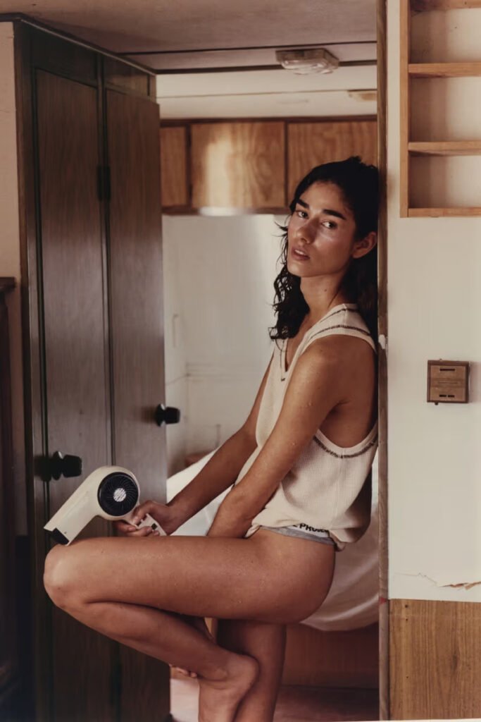 A-model-sits-in-a-vintage-caravan-doorway-holding-a-hair-dryer-wearing-a-cream-waffle-knit-tank-top-and-logo-shorts-from-Nude-Projects-SS26-Airplane-Mode-collection-shot-by-Beltran-Gonzalez