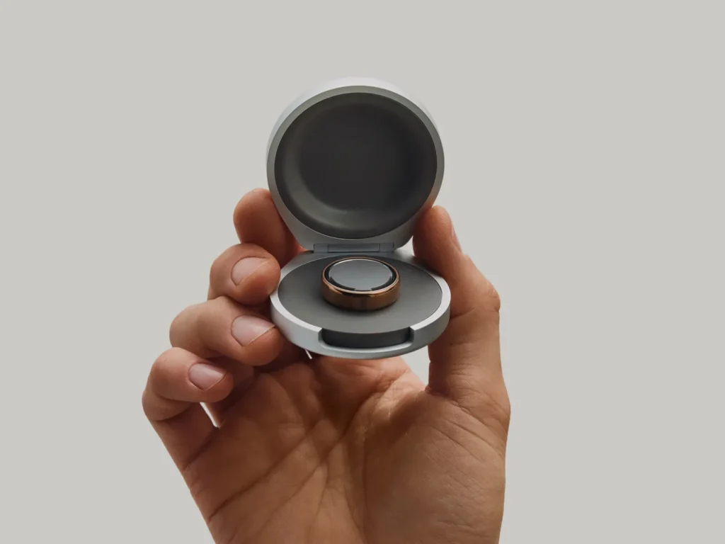 A-hand-holds-the-open-Oura-Ring-5-Charging-Case-in-silver-with-a-gold-Oura-Ring-5-seated-on-the-charging-dock-inside-against-a-neutral-grey-background