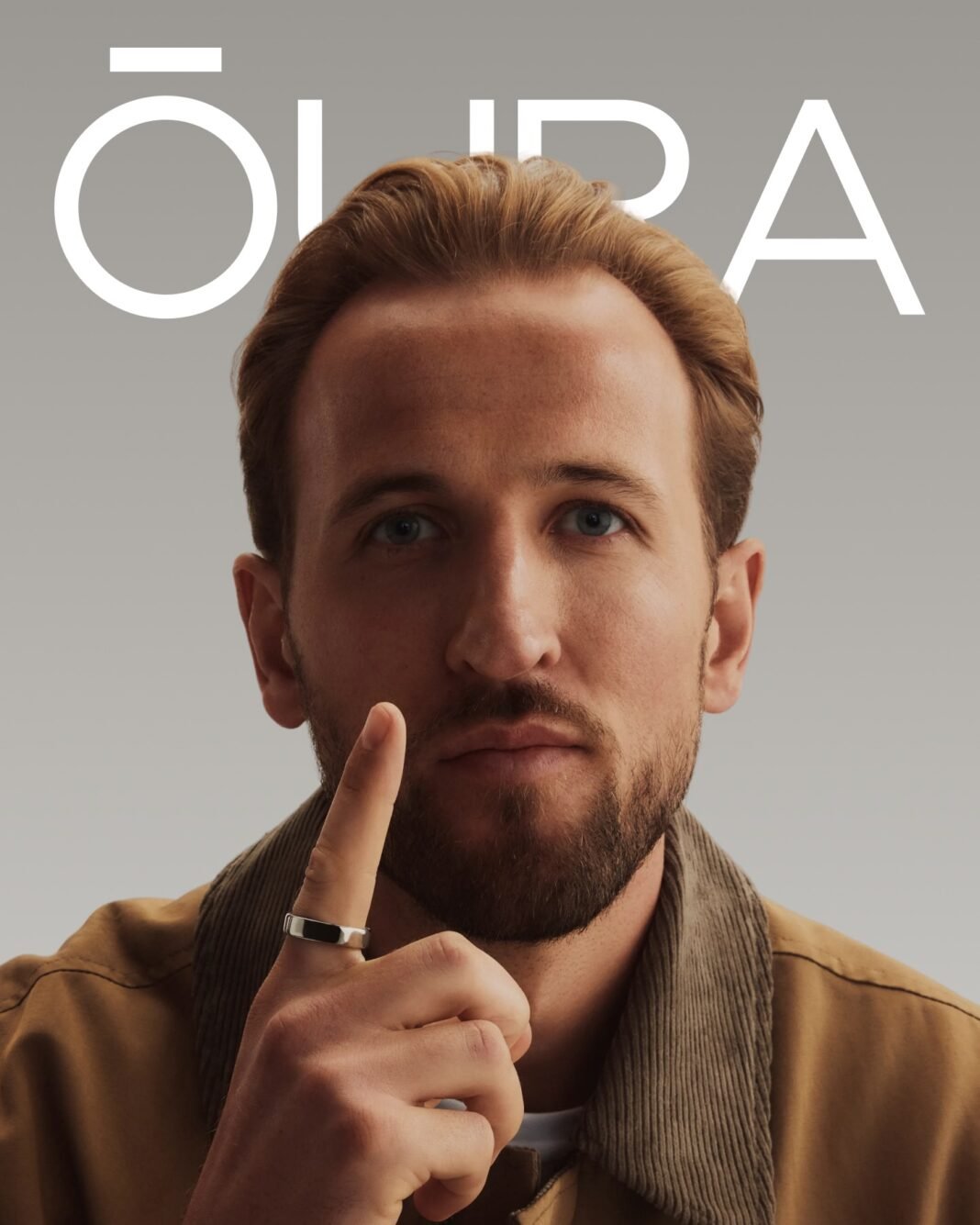 Oura Ring ambassador Harry Kane holds up his hand showing an Oura Ring on his finger, posed against a neutral background with the Oura brand logo overlaid.