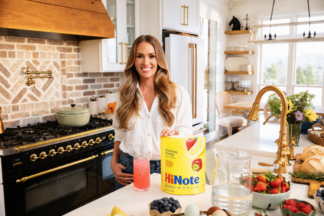 Carrie-Underwood-stands-in-a-farmhouse-kitchen-with-a-yellow-bag-of-HiNote-Everyday-Energy-in-Strawberry-Lemonade-and-a-glass-of-the-pink-drink