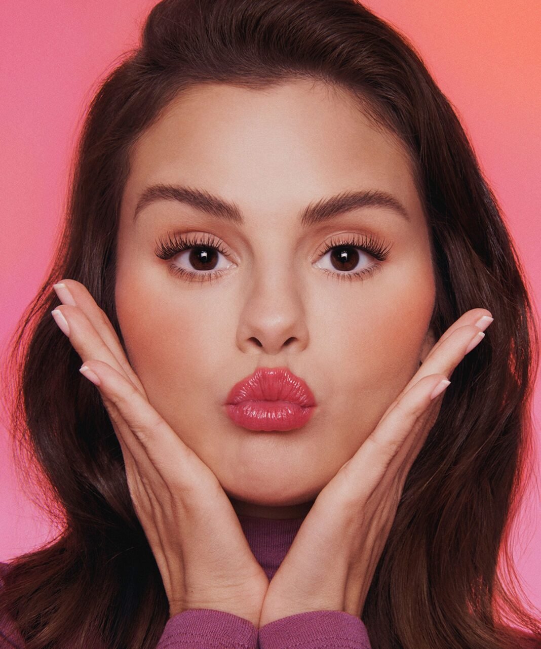 Close-up beauty portrait of a Selena Gomez with long brown hair, wearing glossy coral-pink lipstick and false lashes, pursing her lips in a kiss shape with both hands cupped under her chin.