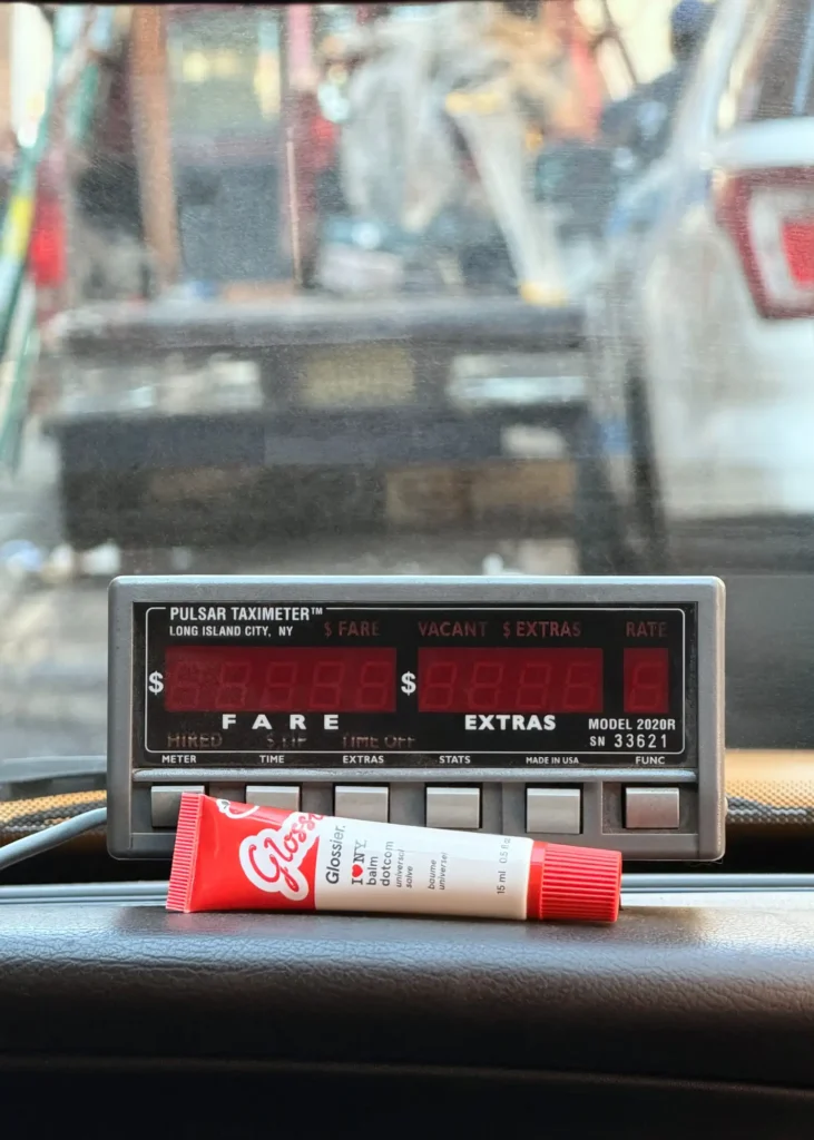 Glossier-I-Love-NY-Balm-Dotcom-tube-resting-on-a-New-York-taxi-dashboard-beside-a-Pulsar-taximeter-city-street-visible-through-the-windshield
