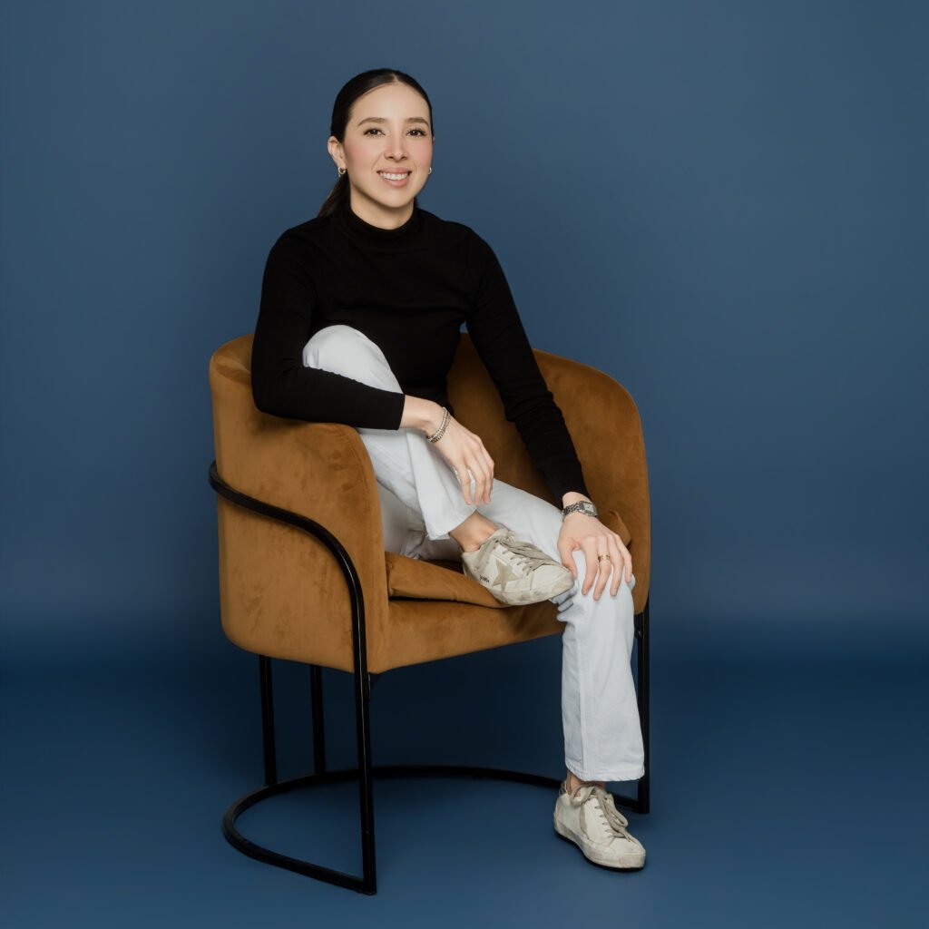 Mariana Maldonado, CMO and CTO of BlueFunnel AI & Co, seated in a tan armchair against a deep blue background, wearing a black turtleneck and white pants