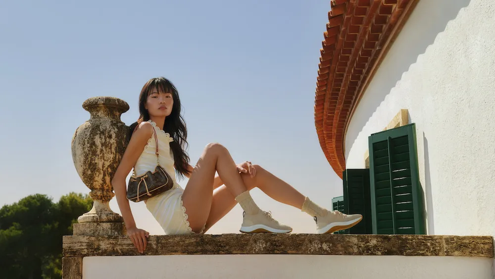 Model-in-a-Louis-Vuitton-scalloped-white-dress-with-a-Monogram-bag-and-trainers-on-a-villa-wall