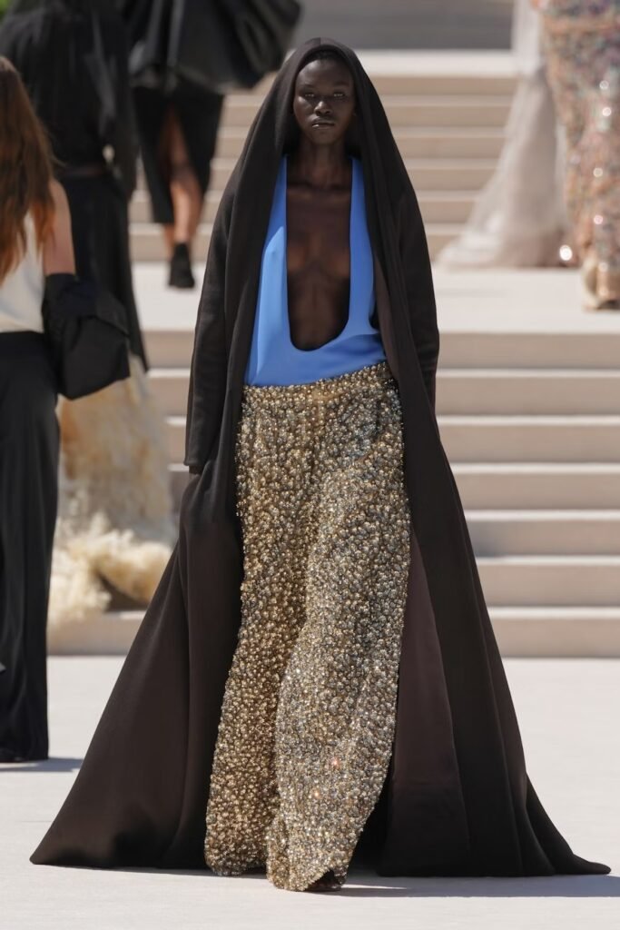 Model-in-a-dark-brown-hooded-cape-over-a-blue-plunging-top-and-gold-beaded-trousers-on-the-Balenciaga-55th-couture-runway-in-Paris