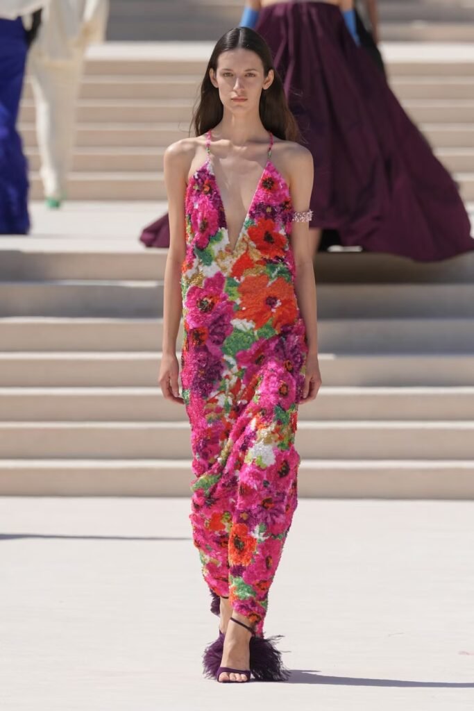 Model in a plunging halter gown covered in textured pink and orange florals with purple feather sandals on the Balenciaga 55th couture runway in Paris.
