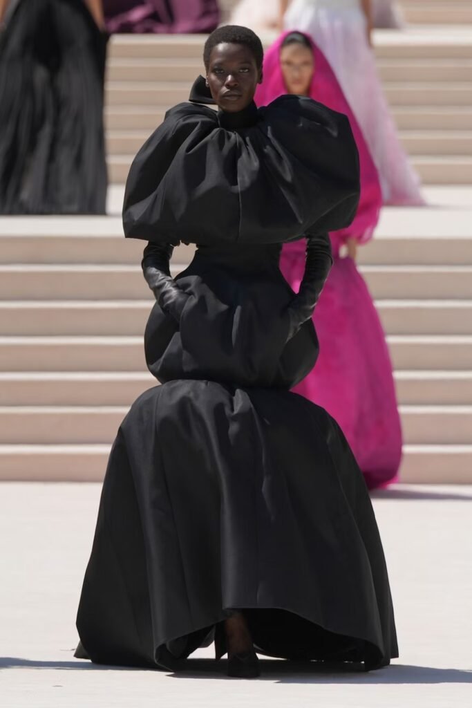 Model-in-a-sculptural-black-gown-with-rounded-balloon-shoulders-and-tiered-volumes-styled-with-black-leather-gloves-on-the-Balenciaga-55th-couture-runway-in-Paris
