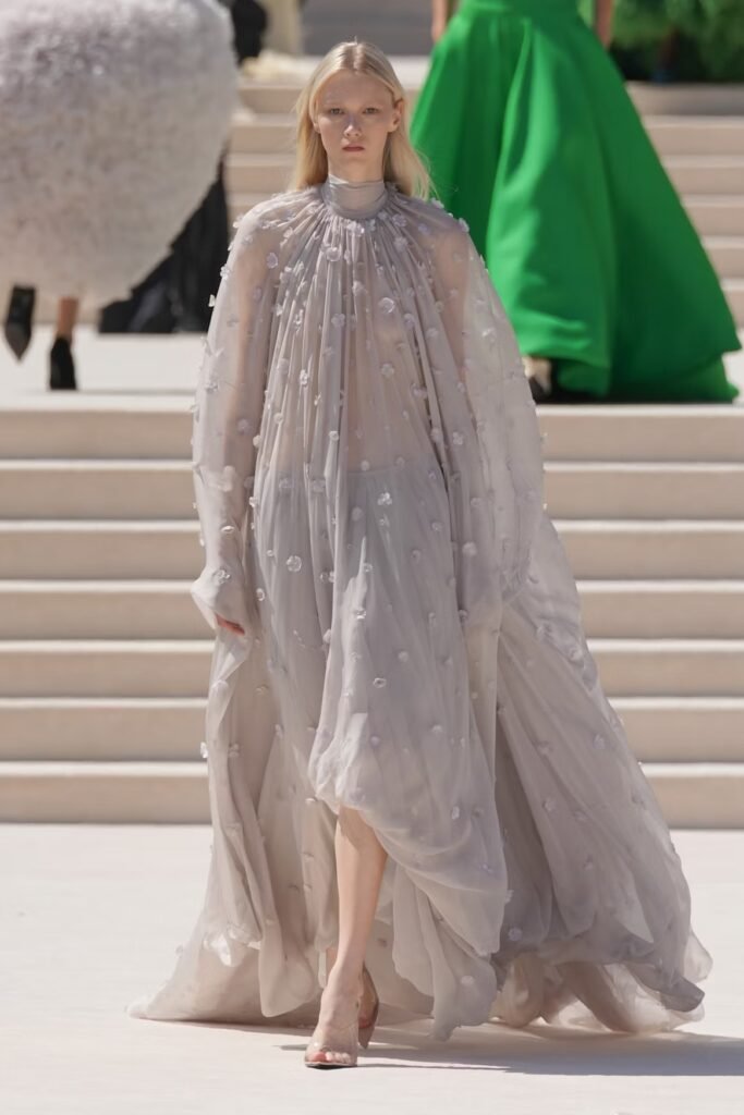 Model in a sheer pale-grey chiffon cape gown with delicate floral appliqués and clear heels on the Balenciaga 55th couture runway in Paris.