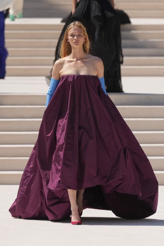 Model in a strapless burgundy taffeta ball gown with blue opera gloves and red pumps on the Balenciaga 55th couture runway in Paris.