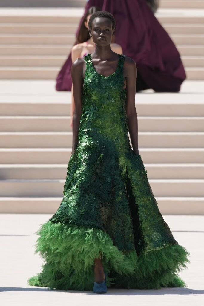 Model in an emerald-green paillette gown with a bright green feather hem and blue heels on the Balenciaga 55th couture runway in Paris.