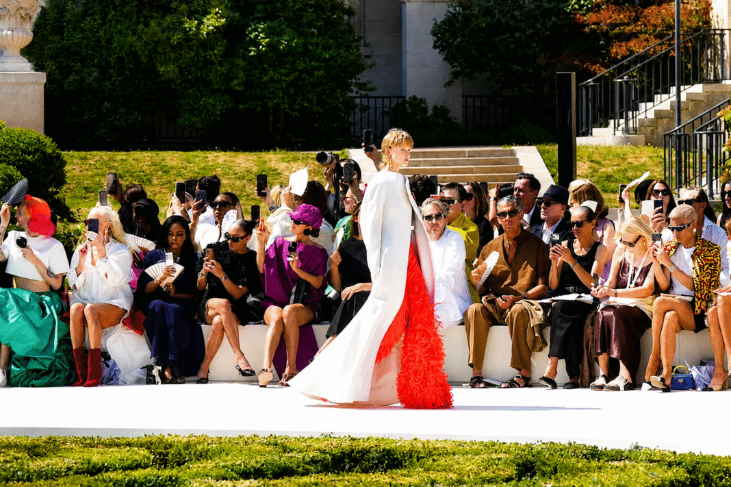 Model in an ivory coat with a scarlet feathered train passing the front row on the Balenciaga 55th couture runway at the Cité Internationale Universitaire de Paris.