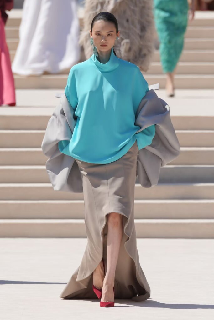 Model-in-an-oversized-turquoise-mock-neck-top-with-a-grey-draped-coat-and-beige-column-skirt-on-the-Balenciaga-55th-couture-runway-in-Paris