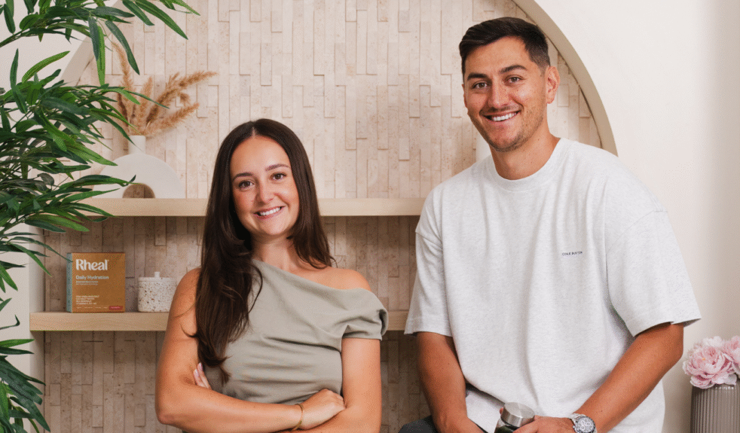 Rheal Superfoods co-founders Charlotte Ali and Sean Ali smiling together beside a shelf displaying a Rheal Daily Hydration box.