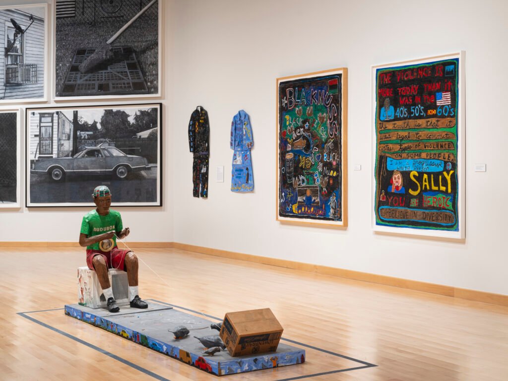 Willie-Birch-sculpture-of-a-boy-trapping-pigeons-with-a-box-near-text-paintings-at-CAAMs-Stories-to-Tell-scaled