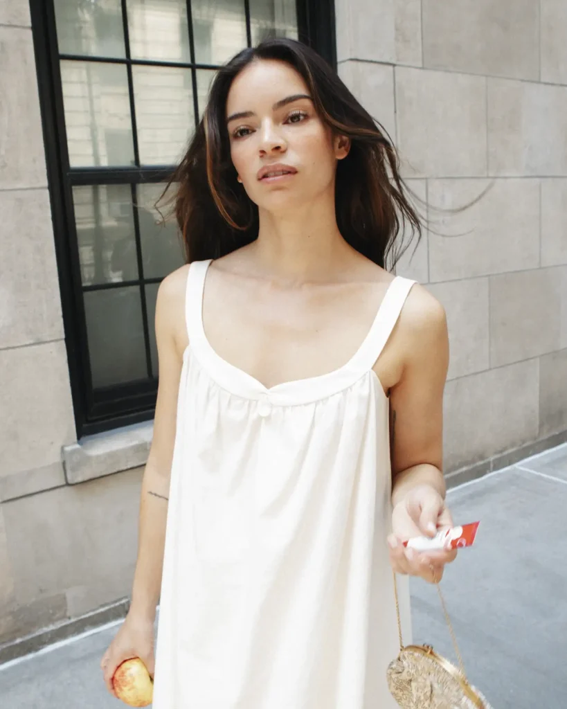 Woman-in-a-white-slip-dress-on-a-New-York-City-sidewalk-holding-a-Glossier-I-Love-NY-Balm-Dotcom-tube-in-one-hand-and-an-apple-in-the-other
