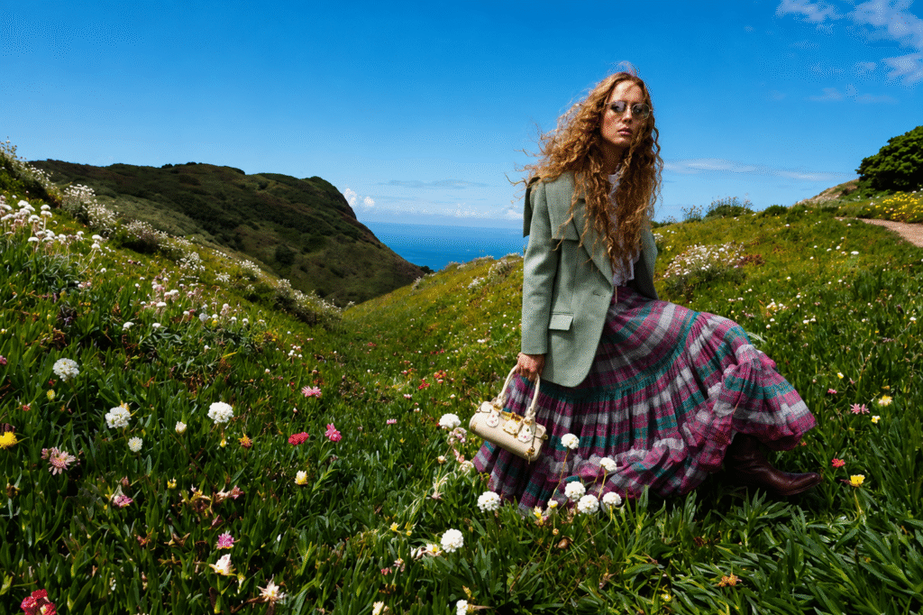 Chloe-campaign-curly-haired-model-in-a-sage-blazer-and-plaid-maxi-skirt-holds-a-cream-bag-on-a-flowering-coastal-hillside