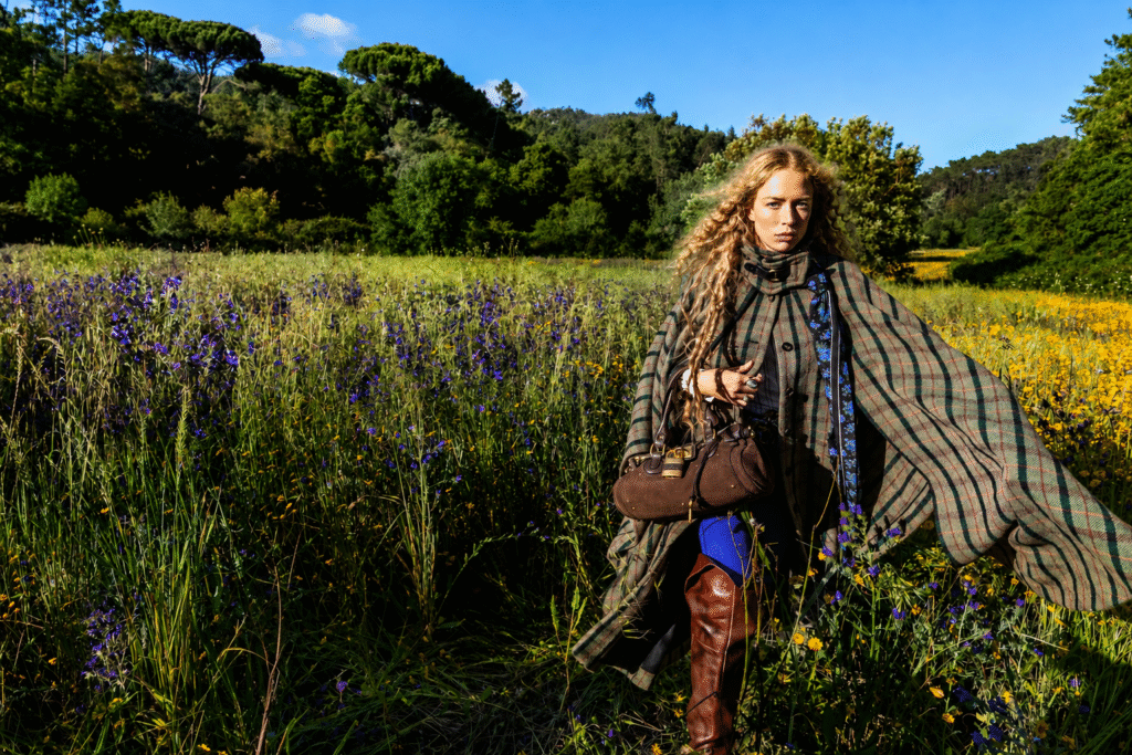 Chloe-campaign-model-in-a-plaid-wool-cape-and-brown-leather-boots-holds-a-suede-bag-in-a-wildflower-meadow