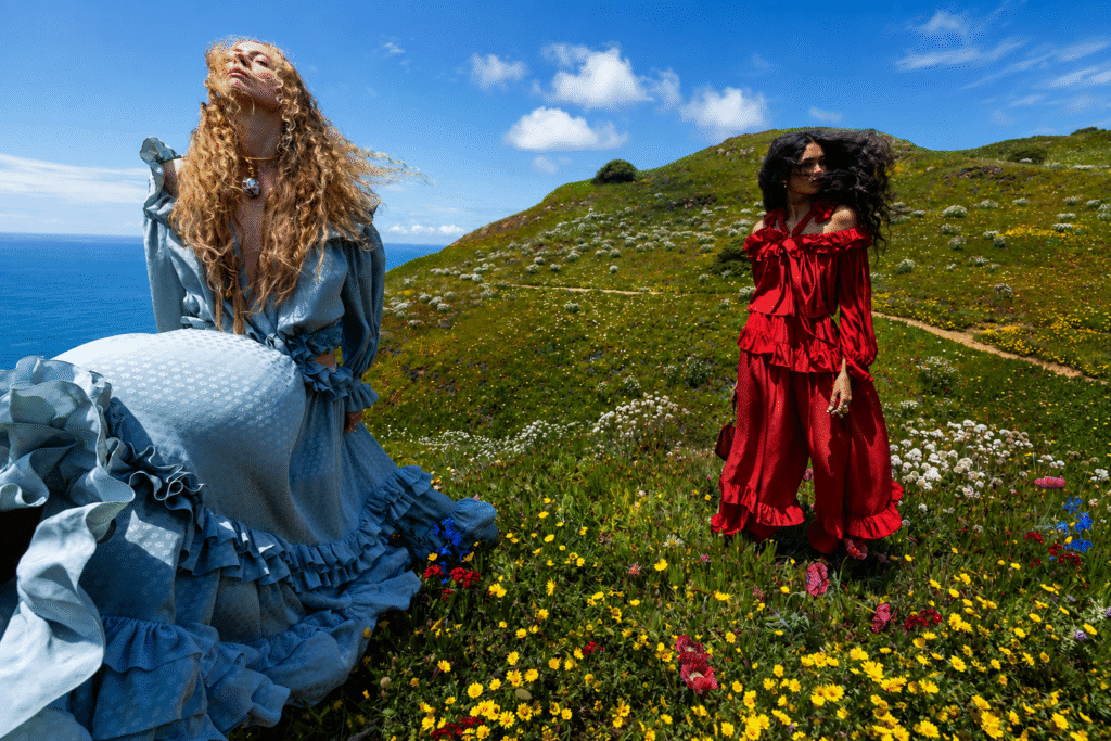 Chloe-campaign-two-models-in-blue-and-red-ruffled-outfits-pose-on-a-wildflower-coastal-hillside-above-the-sea.
