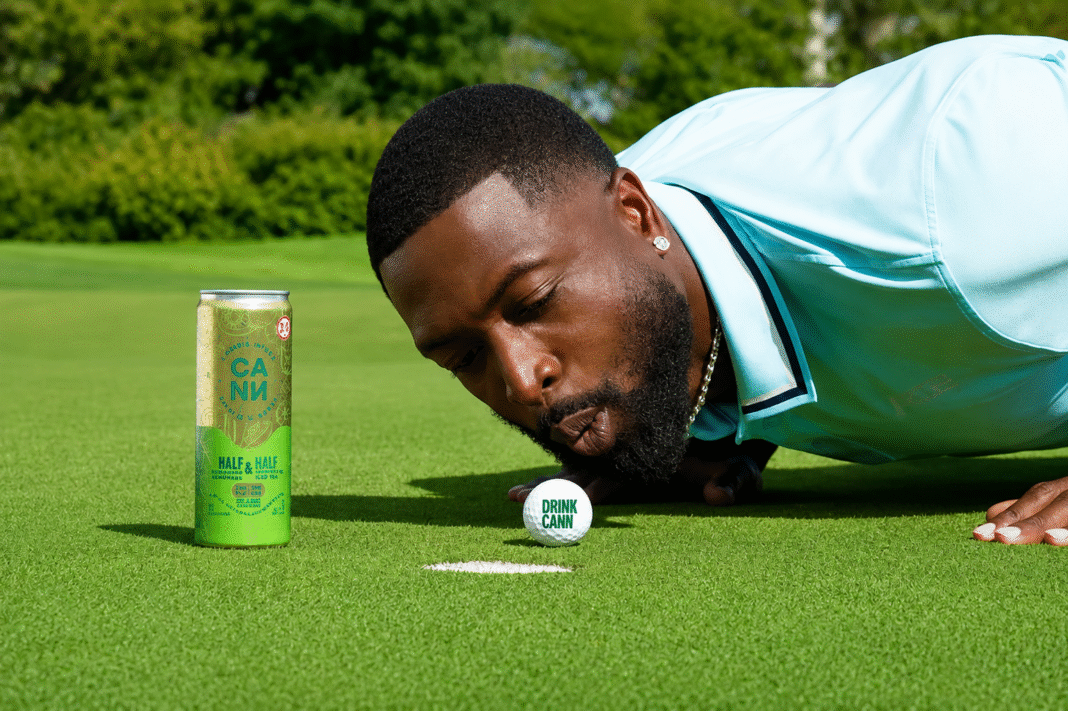 Dwyane Wade in a light blue polo blows a Drink Cann golf ball toward the hole on a putting green beside a Cann Half & Half iced tea can.