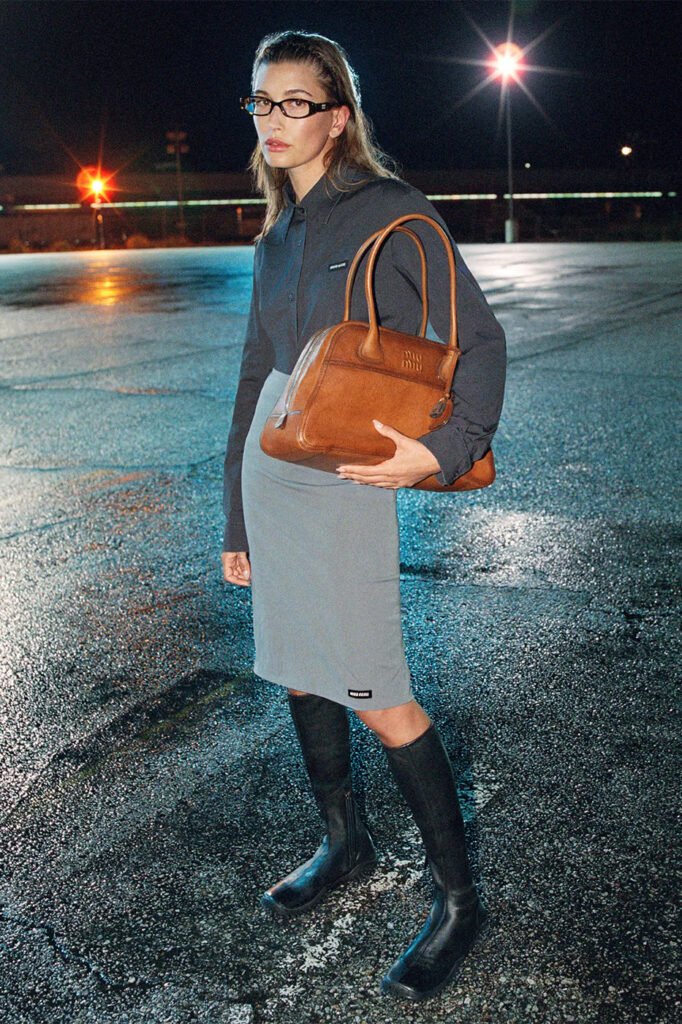 Hailey-Bieber-in-a-grey-shirt-and-pencil-skirt-with-knee-high-black-boots-holding-a-tan-leather-Miu-Miu-bag-photographed-at-night-for-the-Miu-Miu-FallWinter-2026-campaign