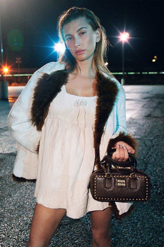 Hailey-Bieber-in-a-white-babydoll-dress-and-fur-trimmed-coat-holding-a-studded-brown-leather-Miu-Miu-bag-photographed-at-night-for-the-Miu-Miu-FallWinter-2026-campaign
