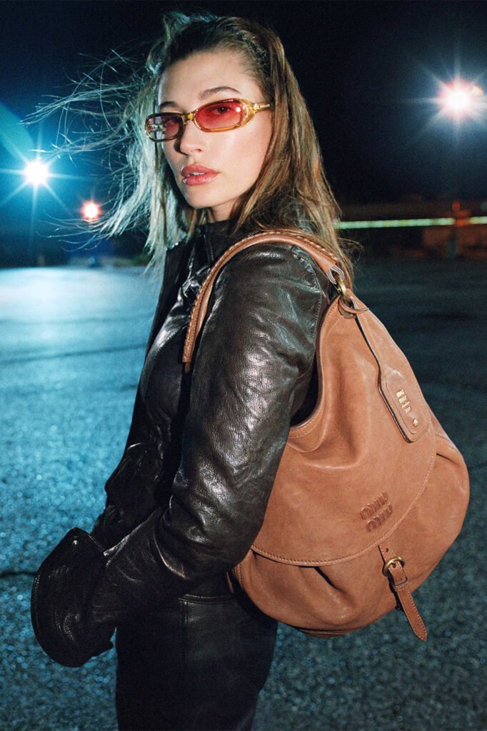 Hailey-Bieber-in-amber-tinted-sunglasses-and-a-black-leather-jacket-carrying-a-tan-suede-Miu-Miu-shoulder-bag-photographed-at-night-for-the-Miu-Miu-FallWinter-2026-campaign