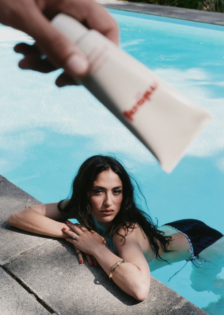 Hand-holding-Misaj-Glow-Protect-cream-tube-above-a-woman-resting-at-a-sunlit-pool-edge