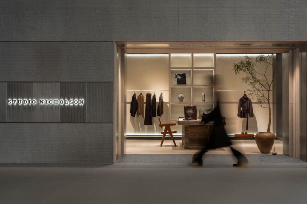 Studio-Nicholson-Beijing-flagship-storefront-at-night-with-the-illuminated-brand-name-on-a-grey-ribbed-facade-and-a-passerby-blurred-in-motion-outside-the-warmly-lit-interior-of-menswear-and-accessories