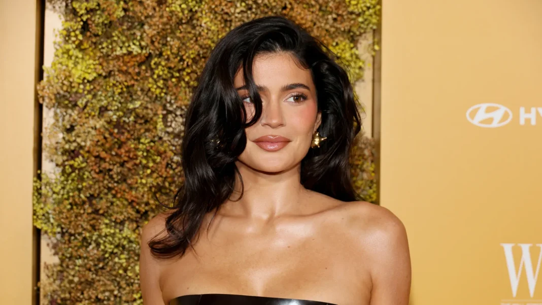 Kylie Jenner in a black strapless top with wavy dark hair and gold earrings, posing before a dried-flower wall.