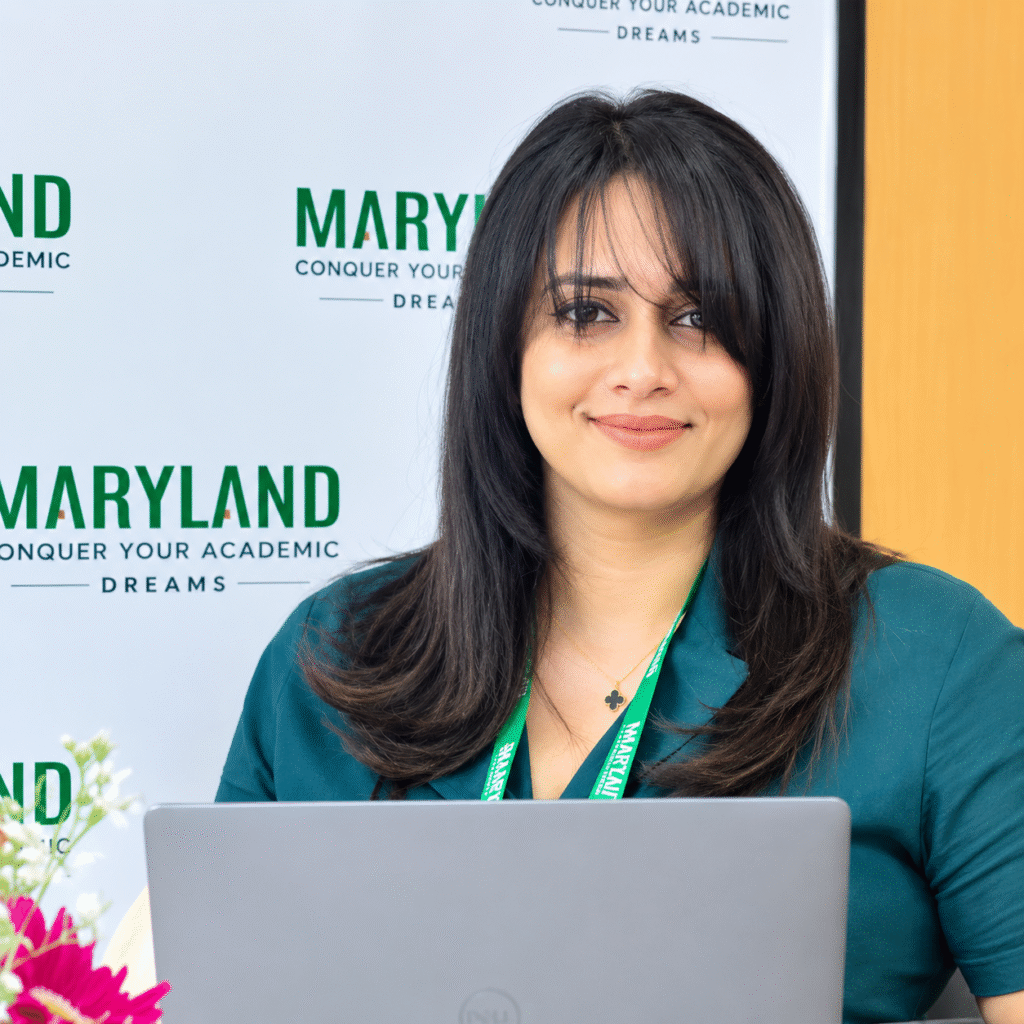 Michelle Morris smiling at her laptop in a teal blouse and Maryland lanyard, in front of a Maryland Consultancy & Academy