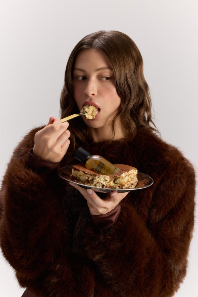 Model-in-a-brown-faux-fur-coat-eating-tiramisu-with-a-DedCool-Mocha-Magic-bottle-resting-on-the-plate
