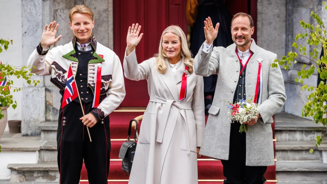 Queen Mette-Marit waves in a grey coat with an oxygen cannula, flanked by Prince Sverre Magnus and King Haakon VIII in bunad.