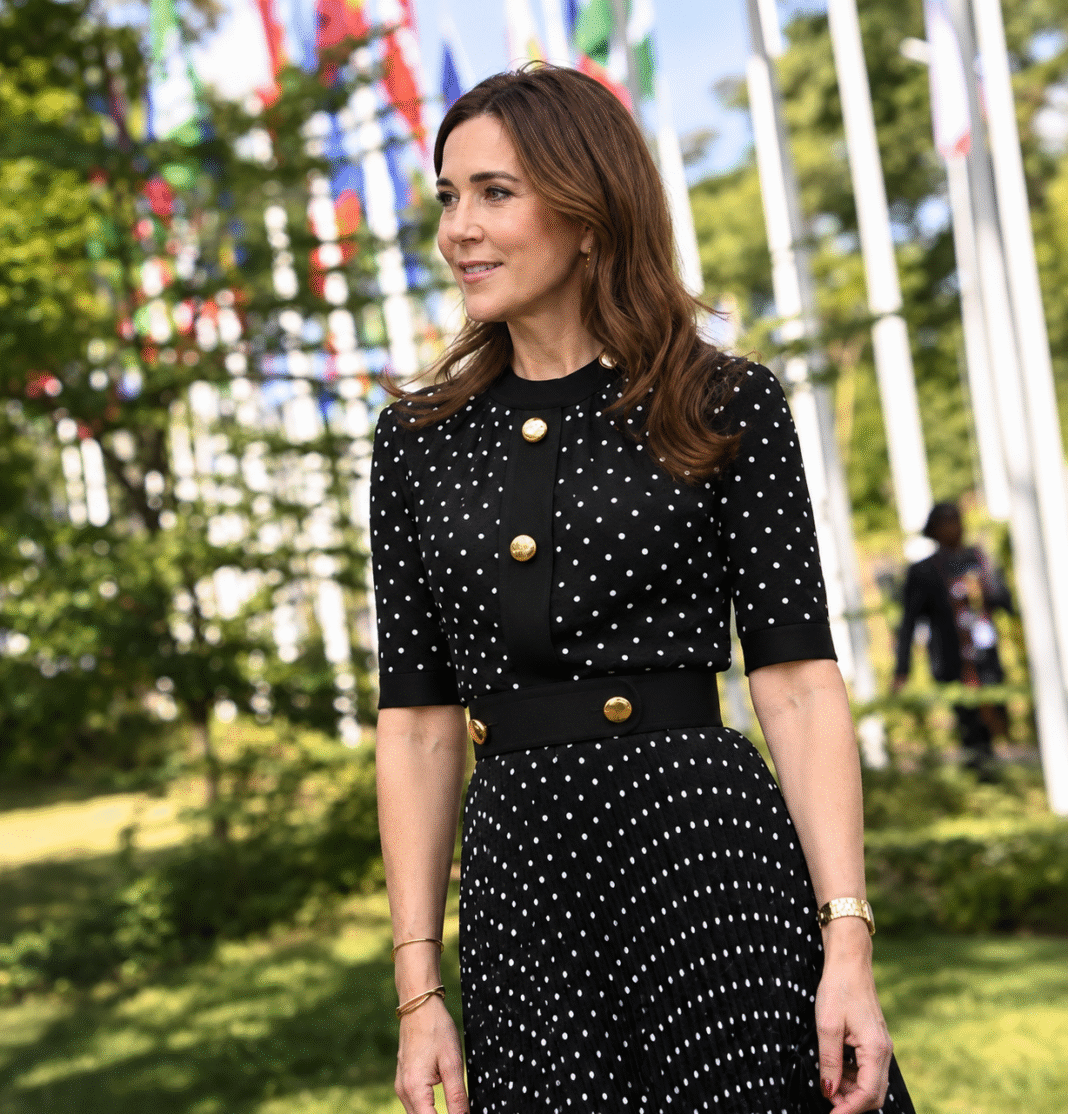 Queen Mary of Denmark smiles outdoors in a black polka dot dress with gold buttons in front of a row of international flags on tall flagpoles.png