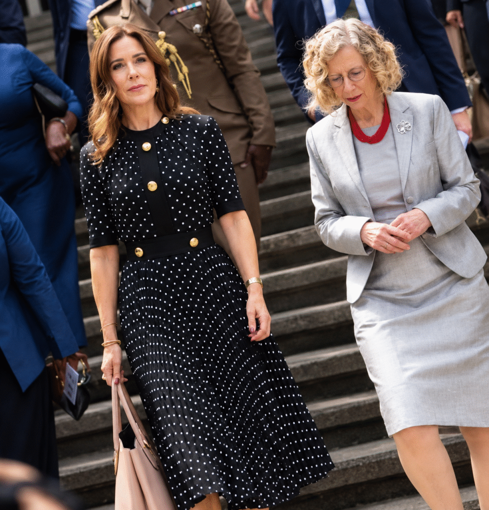 Queen Mary of Denmark walks down stone steps in a black polka-dot midi dress with gold buttons, a pleated skirt and a blush tote.