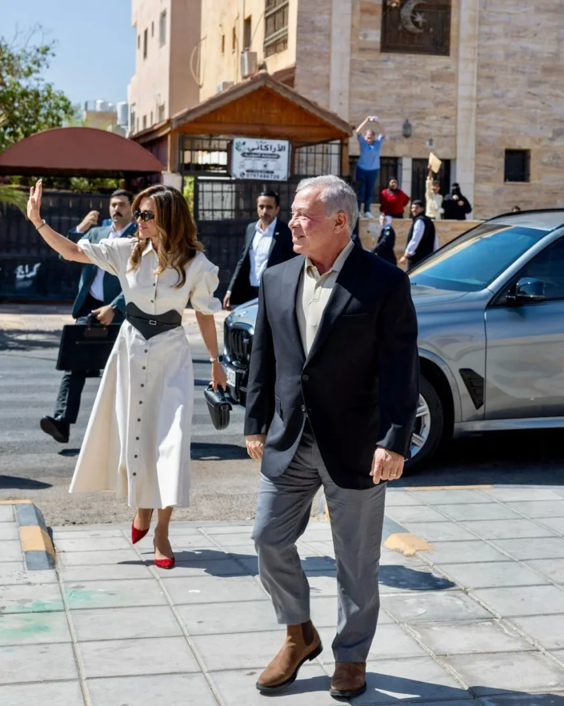Queen Rania in an ivory puff-sleeve shirtdress with a black corset belt beside King Abdullah II at a school in Aqaba.